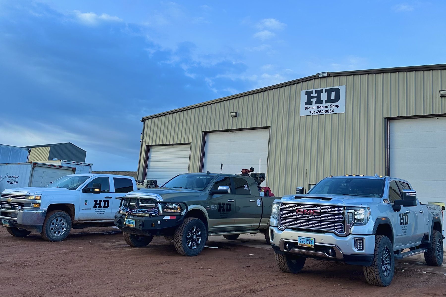 Three pickups lettered HD parked along the shop wall under a wide blue North Dakota sky