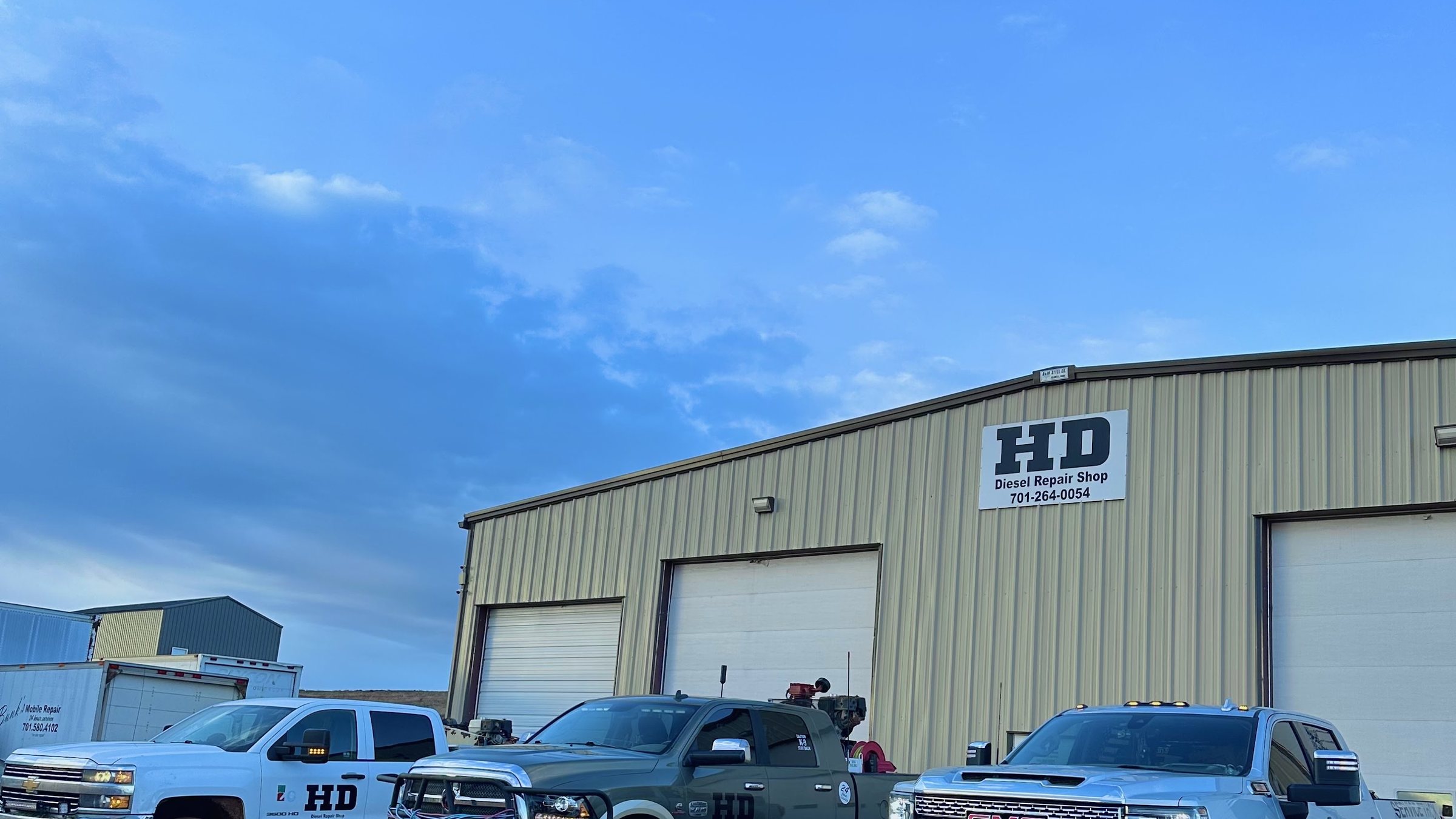 The HD Diesel shop in Killdeer, North Dakota — a long steel building with the HD sign over the doors and the crew's pickups parked along the front under a wide blue sky