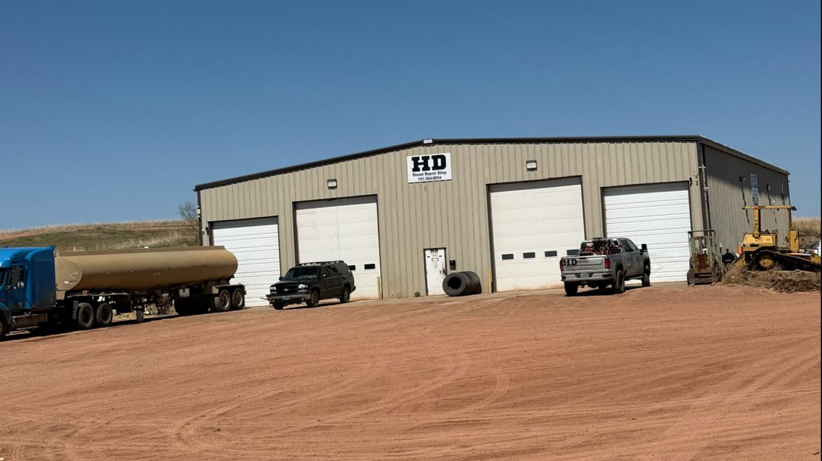 The HD Diesel shop in Killdeer — a long tan steel building with four overhead doors, a tanker trailer and pickups parked on the red dirt lot